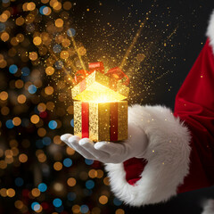 Close-Up of Santa Claus's Hand Holding an Opened Golden Gift Box Exploding with Bright Magical Light and Sparkling Particles, Set Against a Background of Defocused Christmas Tree Lights