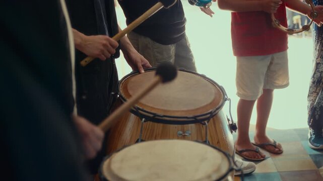 Close-up of people playing drums and tambourines in a group, showing hands, percussion instruments, and a bright indoor setting