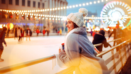 A woman smiles while leaning on the railing of an ice skating rink surrounded by twinkling lights....