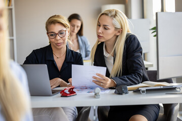 Women collaborating at a modern office, discussing documents and working on a laptop during a...