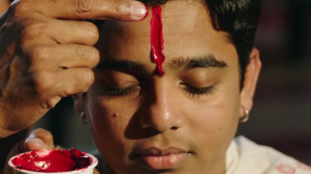 Close up shot of an adult hand applying sacred red tilak mark onto a young boy forehead during a spiritual ritual
