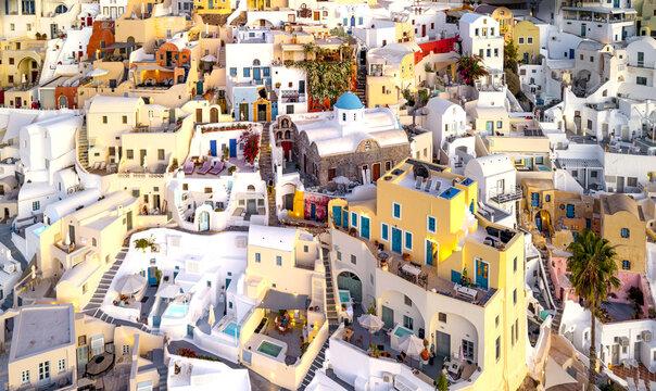 Aerial view of white-washed buildings cascade down the cliffs, punctuated by blue domes and vibrant bougainvillea, creating a mesmerizing tapestry of Cycladic charm, Oia, Thira, Greece.