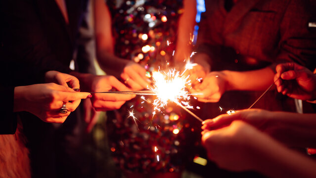 Hands hold sparklers, creating bright sparks in a lively setting. Friends celebrate together as lights twinkle and laughter fills the air during a special event.
