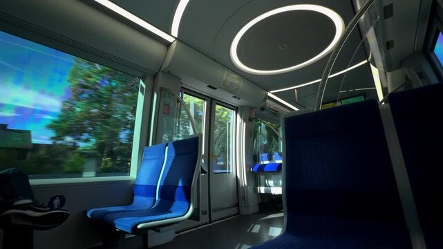 Innenansicht eines modernen Muenchener U-Bahn-Wagens mit hellen, runden Lichtern und blauen Sitzen. Durch das Fenster faehrt die U-Bahn oberirdisch durch die sonnige Stadtlandschaft