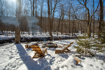 Snowy woodland fire pit with wooden chairs around a small flame beside a creek on a bright winter day. Peaceful outdoor nature setting.