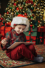 A child wearing a Santa hat and a plaid shirt with a bow tie writes a letter to Santa under the...