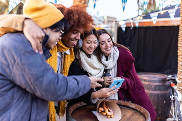Group of multi-ethnic friends looking at photos on a smartphone at christmas market, hugging,...