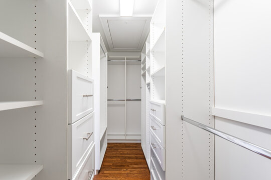 Bright walk-in closet with built-in white shelving, drawers, and hanging space. Clean, organized modern storage interior with hardwood flooring. - Powered by Adobe