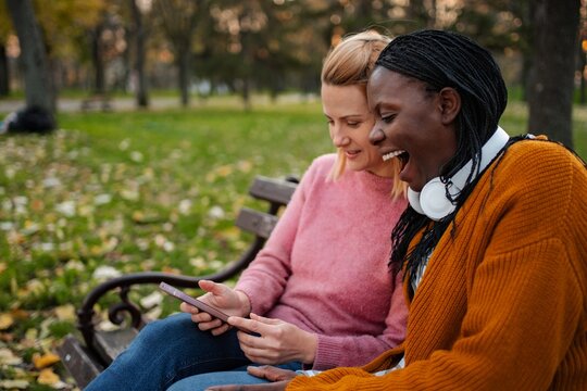 Diverse women friends laughing watching smartphone video on park bench
