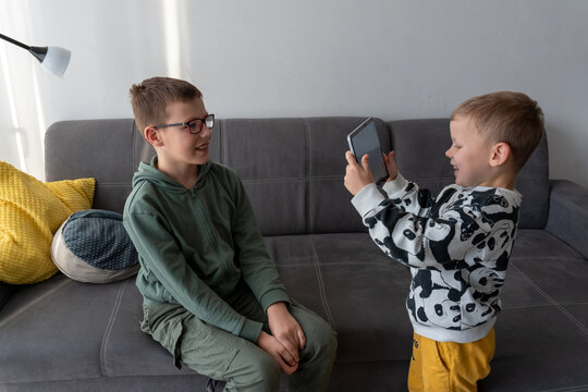 Little boy taking photo of smiling older brother with tablet while standing in living room at home. Concept of siblings creative play, kids photography fun and childhood digital skills development.