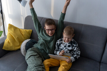 Excited boy with raised arms celebrating while younger brother holds tablet on grey sofa at home. Concept of gaming victory, siblings digital fun, childhood excitement, screen time entertainment joy.