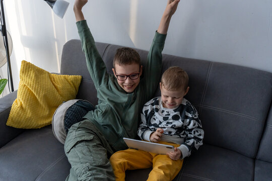 Excited boy with raised arms celebrating while younger brother holds tablet on grey sofa at home. Concept of gaming victory, siblings digital fun, childhood excitement, screen time entertainment joy.