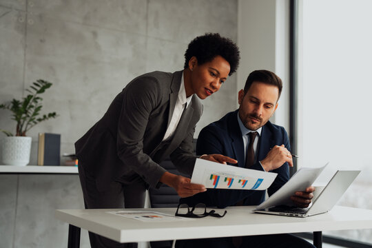 A businessman and an African businesswoman discussing a project in a modern office while using a laptop. They appear focused and engaged in a professional conversation