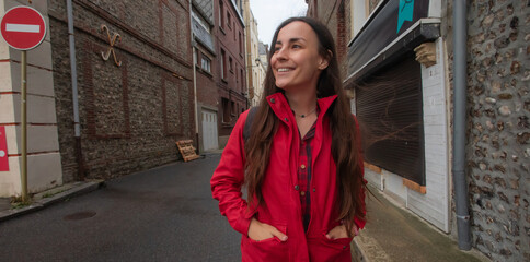 Smiling young woman in a red jacket walking through a narrow European street, hands in pockets, enjoying an urban stroll. Casual travel mood in an old town alleyway.