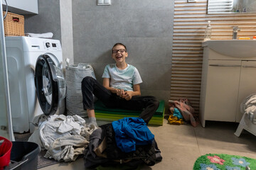 Happy laughing boy sitting on floor near washing machine surrounded by piles of clothes in bathroom at home. Concept of fun household chores, making laundry enjoyable and positive childhood memories.