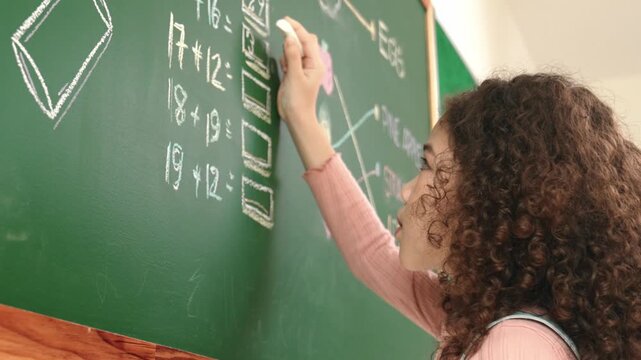 Back view of african student answer question while writing at board. Teacher looking at elementary student doing math quiz in front of classroom while smart children attend in math lesson. Pedagogy.