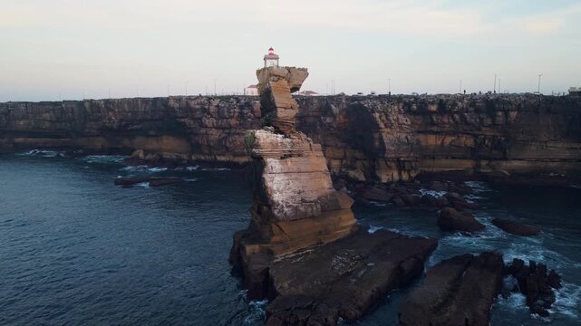 Aerial view of Cabo Carvoeiro sea stack and towering stratified cliffs with lighthouse in the background as waves crash against the rugged Atlantic coastline in soft evening light