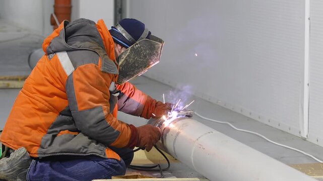 Welder welding a metal pipe or pipeline. Close-up of welding work indoor. Welder at a construction site. Welding work inside a large industrial or commercial building. Welding seam, sparks, flash.