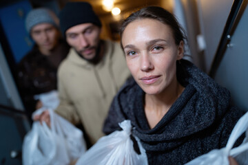 Three people, with one woman in the foreground, hold grocery bags while navigating a stairway, showcasing a candid moment of domestic life and shared responsibilities.