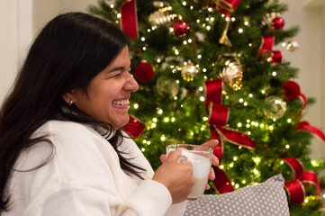 Smiling Woman Sitting by Decorated Christmas Tree. Woman Relaxing With Hot Drink by Christmas Tree.