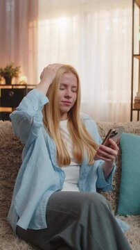 Young woman sits at home holding smartphone, discovering bad news and feeling shock, sadness and regret. Caucasian girl collapses on couch, revealing despair after phone defeat fail under sunset glow
