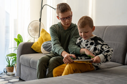 Older brother teaching younger sibling to play on tablet while sitting together on grey sofa in bright living room. Concept of siblings mentoring, digital learning and brotherly bonding moments. - Powered by Adobe
