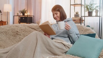 Young woman relaxing reading interesting book enjoying literature while having cup of hot chocolate, cocoa or tea on comfortable sofa. Smiling girl wrapped in a warm blanket in living room at home. - Powered by Adobe