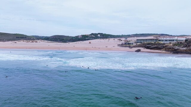 Aerial wide shot of surfers in water and people walking along sandy dunes at Guincho Beach, Portugal with ocean waves and coastal buildings