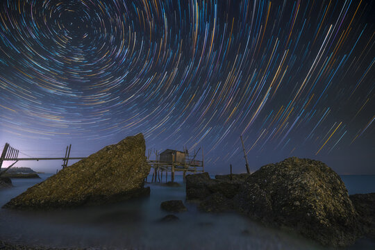 View of star trails streak across the inky sky above the tranquil sea, illuminating the ancient trabucco perched on stilts, a dance of time and tradition, Chieti, Abruzzo, Italy.