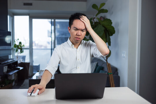 Young man sitting at home with a laptop, holding his head in frustration while feeling stuck on a task. Captures remote work stress and problem-solving struggle in a home office environment.