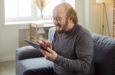 Happy senior, old mature man using computer tablet at home, looking at screen, laughing at funny joke or video in social network, holding new electronic device, sitting on couch, learning to use