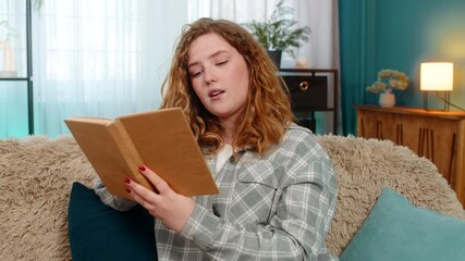 Young woman at home reading book with focus and flipping pages slowly while reflecting on deep content. Caucasian redhead girl on sofa engaged in thoughtful reading, enjoying quiet intellectual moment - Powered by Adobe