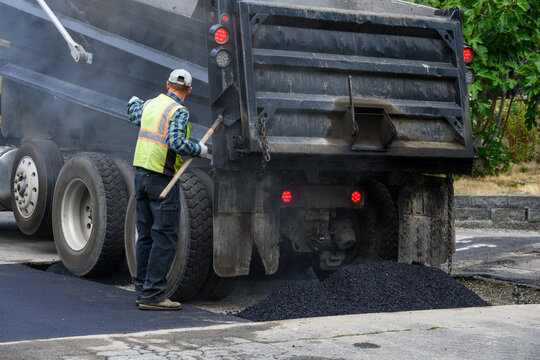 Construction worker on roadworks paving repair project with shovel directing truck driver to hold on dumping hot asphalt from dump truck
