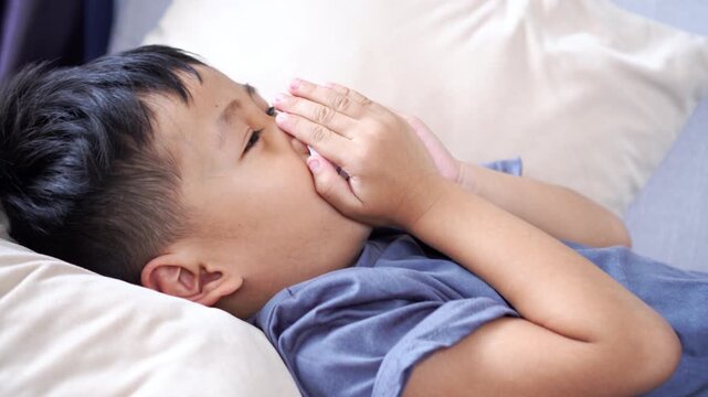 Asian boy sneezing, coughing, and suffering from a runny nose while sick at home, wiping his nose with a tissue and showing clear symptoms of a cold, flu, and respiratory infection.