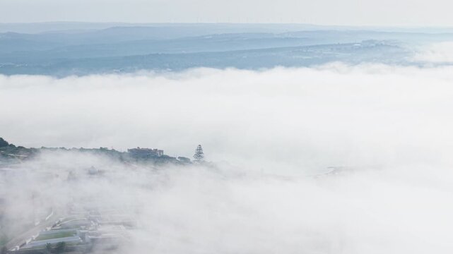 Aerial view of rolling hills and village rooftops emerging through thick morning fog with layered landscape in the distance