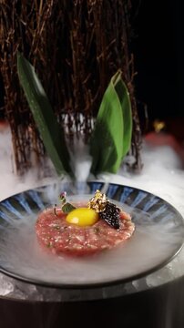 Luxury beef tartare with egg yolk, caviar and edible gold leaf on elegant plate, dramatic smoke effect and cinematic rightward camera movement in upscale fine dining restaurant
