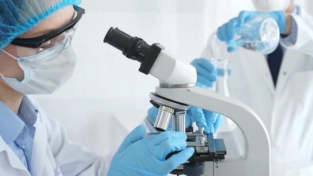 Female scientist wearing protective gear is examining specimen under microscope while man laboratory assistant handles glassware in a professional medical investigation. Medicine and science concept