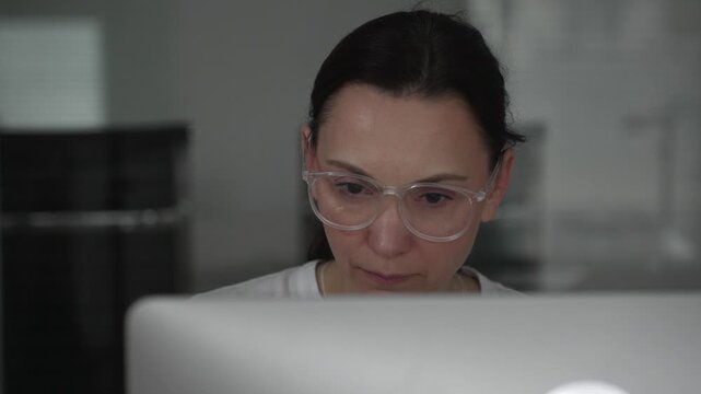 Woman wearing clear frame glasses intently focused on a computer screen in a modern office, conveying concentration, professionalism and engaged digital work. Business people concept