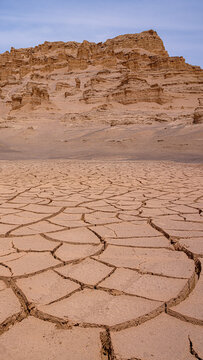 Aerial view of parched earth fractures into geometric patterns under a pale sky, with the towering desert landscape of the Lut Desert rising in the distance, Shahdad, Kerman Province, Iran.