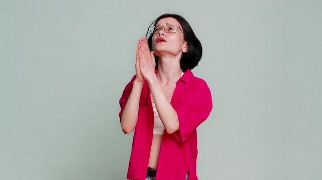 Young woman gazes upward holds hands together in prayer, emotionally asking God for help in difficult situation. Caucasian girl expressing sincere hopeful plea and faith, isolated on gray background.