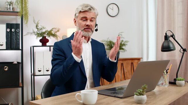 Middle-aged smiling businessman at home office wears headset assisting hotline caller via laptop. Freelancer guy at table gives rapid support ensuring customer satisfaction enhancing service quality