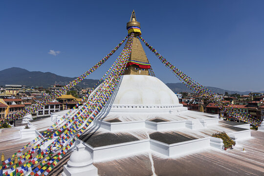 View of the Boudhanath Stupa's majestic white dome rises, adorned with prayer flags fluttering in the breeze against a clear blue sky, Boudhha, Bagmati Province, Nepal.