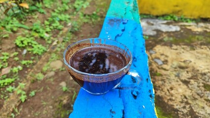 A clear glass cup of black coffee sits on a vibrant blue wooden surface outdoors, with a natural green background creating a calm and rustic atmosphere
