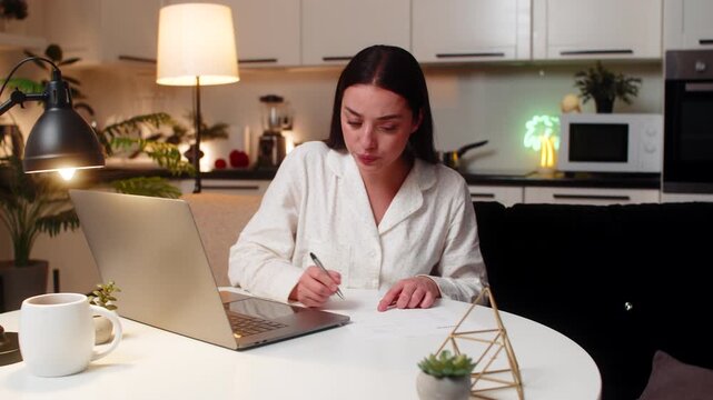 Young woman at night home sees large utility bill holds receipt frowns feels stress anger wants fix. Caucasian girl on sofa at evening throws papers sighs plans dispute calls support to check error.