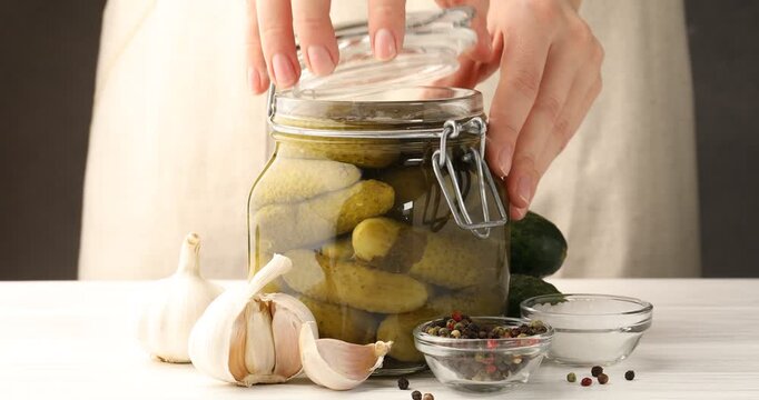 Woman opening jar and taking pickled cucumber at white wooden table, closeup