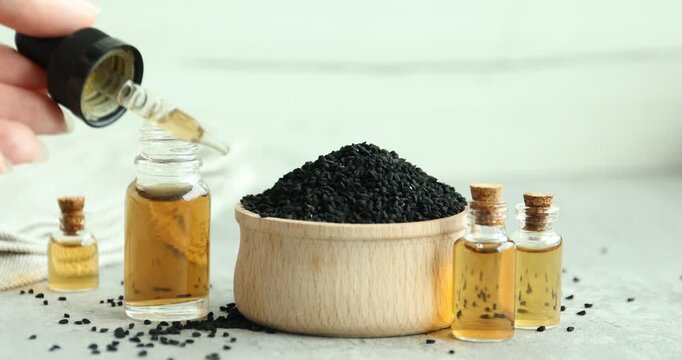 Woman taking pipette from bottle of essential oil and putting it onto grey table with black cumin seeds, closeup