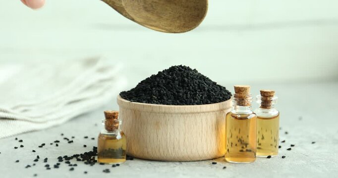 Woman adding black cumin seeds from spoon into bowl at grey table with bottles of essential oil, closeup