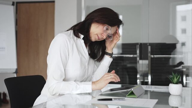 Businesswoman in white blouse and glasses sitting at a modern desk, working with a tablet in a contemporary office, focusing on digital tasks, using technology for efficient workflow