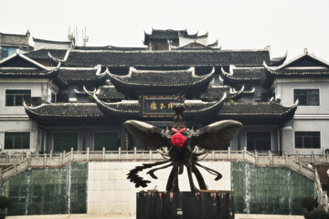 View of dark, traditional architecture looms behind a sculpture with a touch of vibrant red, creating a striking contrast, Xiangxi Tujia, Hunan, China.