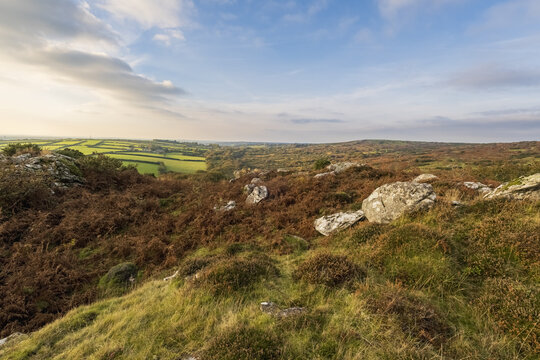 View of rugged, rocky terrain covered in brown bracken and green moss, contrasting with distant patchwork fields under a vast sky, Bodmin, England, United Kingdom.